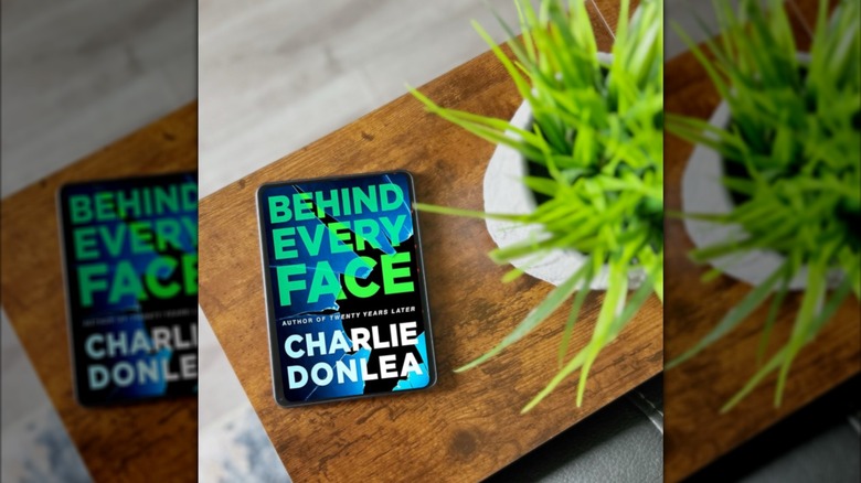 A copy of "Behind Every Face" laying on a table next to a plant