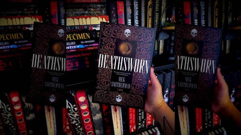 A hand holding up a copy of "Deathsworn" in front of bookshelves