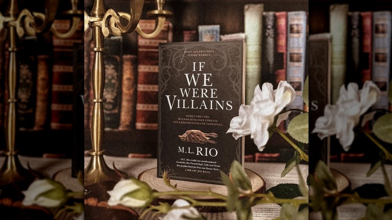 A copy of "If We Were Villains" sitting on a table surrounded by plants and old books