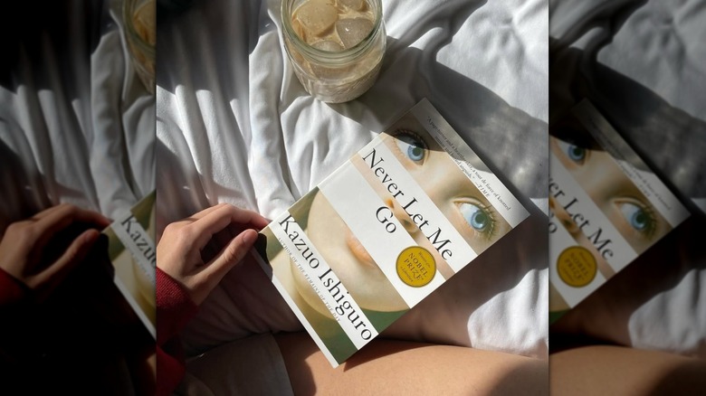 A copy of the novel "Never Let Me Go" sitting on a bed next to a woman's leg and a cup of iced coffee
