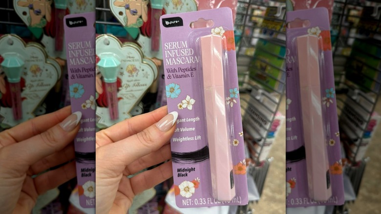 Hand holding up the b.pure Serum Infused Mascara, still in the package, with a cosmetics display in the background