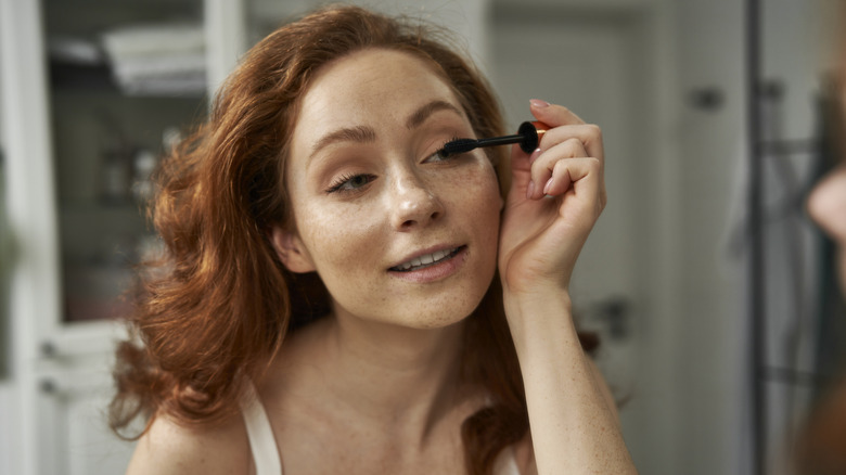 Freckled red-head applying mascara in a bronze tub to her eyelashes