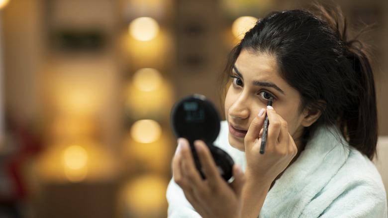 A young woman applying eyeliner while in a white bath robe
