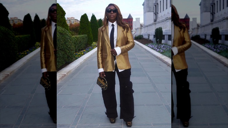 A woman wearing a shirt and tie under a gold blazer and black pants