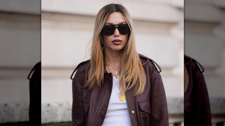 Woman with blond hair and darker roots with a center part, oversized black square sunglasses with dark lenses, a burgundy leather bomber jacket, a white ribbed tank top