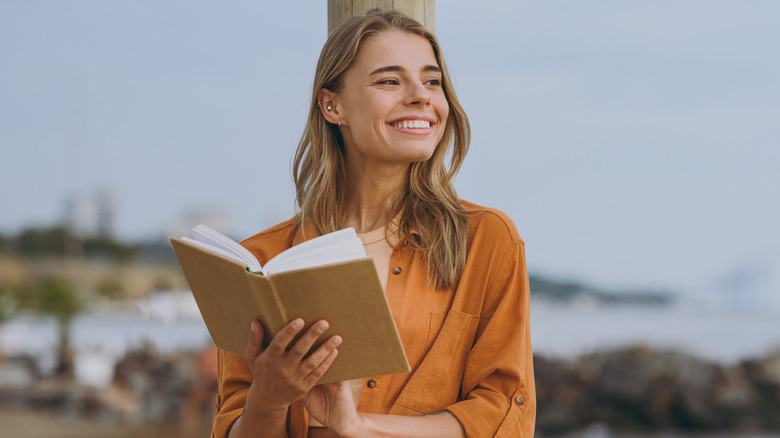 A woman holding a book and standing on a beach reading