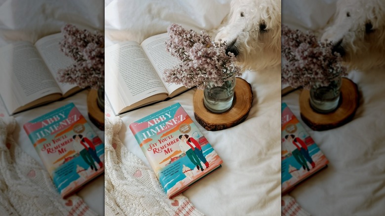 A copy of Say You'll Remember Me laying on a bed with flowers and a dog