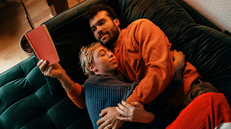 A couple lies on a sofa holding each other as one reads