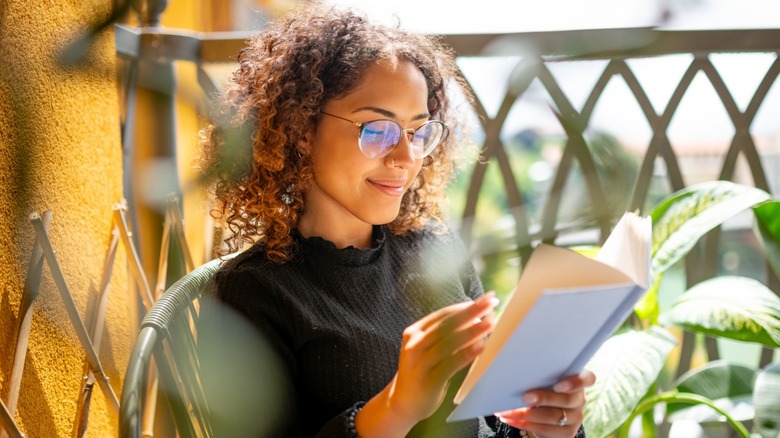 A woman with glasses reading a blue book while on a balcony