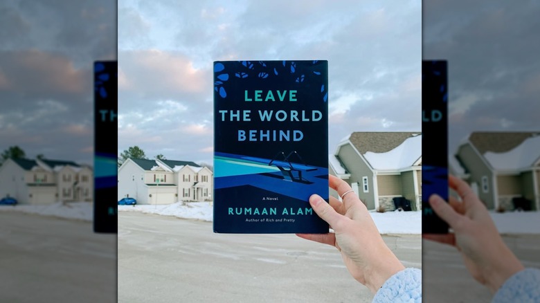 A hand holding a copy of the novel "Leave the World Behind" in front of a house and overcast sky