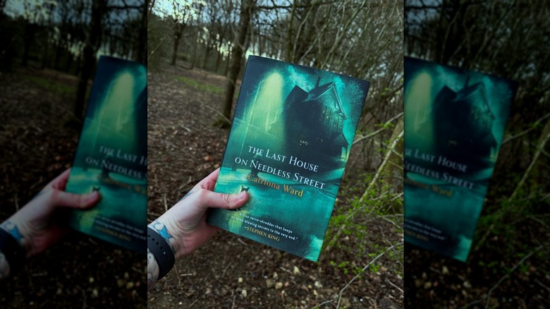 A hand holding a copy of the novel "The Last House on Needless Street" with woods in the background