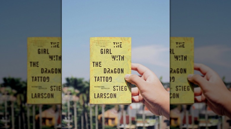 A hand holding a copy of the novel "The Girl with the Dragon Tattoo" in front of a blue sky