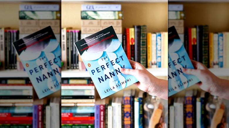A hand holding a copy of the novel "The Perfect Nanny" in front of a full bookshelf