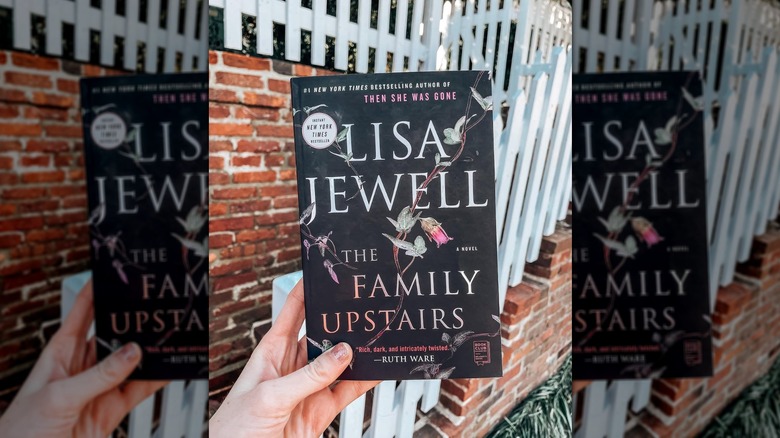 A hand holding a copy of the novel "The Family Upstairs" in front of a brick wall