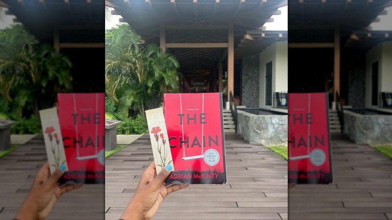 A hand holding a copy of the novel "The Chain" in front of a house