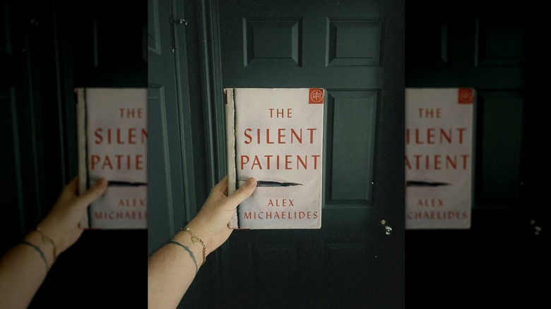 A hand holding up a copy of the book "The Silent Patient" in front of a closed door