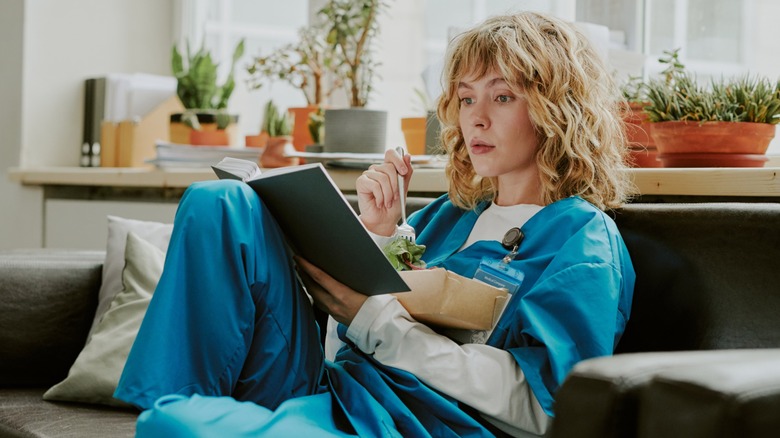 A woman reading a book while eating a salad and wearing scrubs