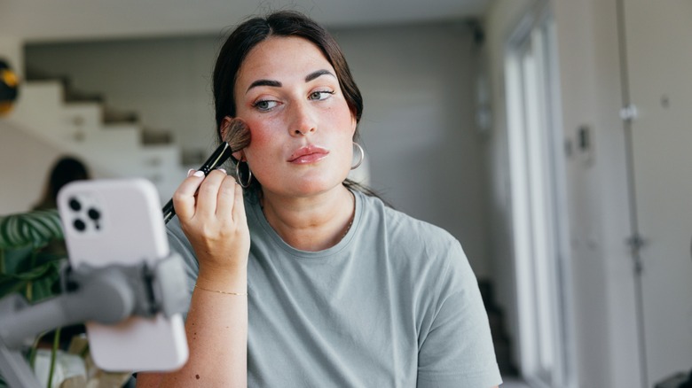 A woman applying blush to her cheeks while recording herself with a phone