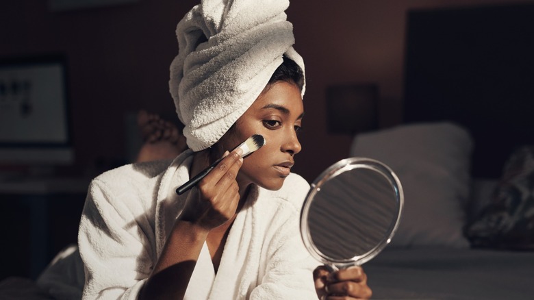 A woman looking in the mirror and applying foundation with a brush