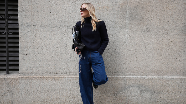 A person wearing a black sweater and dark blue barrel jeans, leaning against a wall with one leg resting on the wall behind them