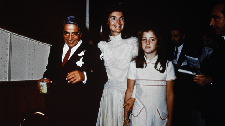 Jacqui Kennedy in a white shift dress next to a young Caroline Kennedy and Aristotle Onassis