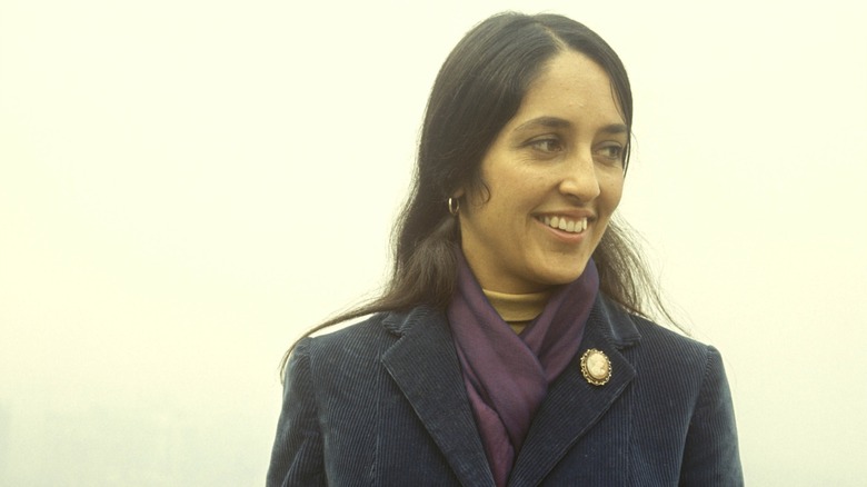 Joan Baez smiling outside in a purple coat and scarf in 1960