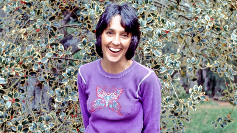 Joan Baez smiling while standing in front of a plant in 1972