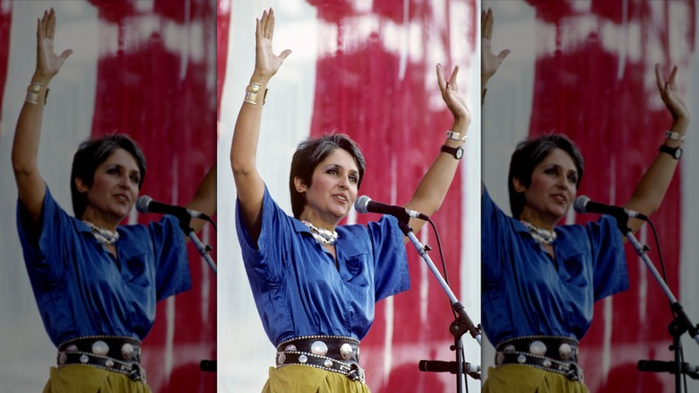 Joan Baez with her arms raised while wearing a blue top and yellow bottoms while on stage performing at Live Aid 1985