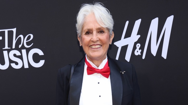 Joan Baez smiling on the red carpet in 2023 while posing in front of a black background wearing a red bowtie