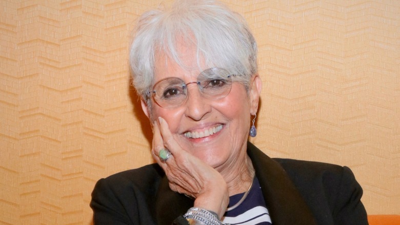 Joan Baez smiling with her hand on her face in front of an orange background