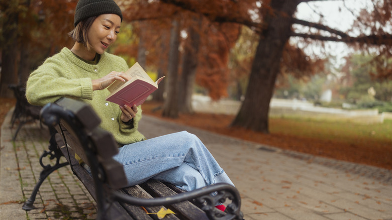 Woman with short hair and black beanie reading while sitting on a park bench with an autumnal landscape