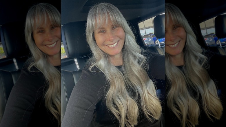 An older woman smiling in a car selfie with long, greying hair and bangs