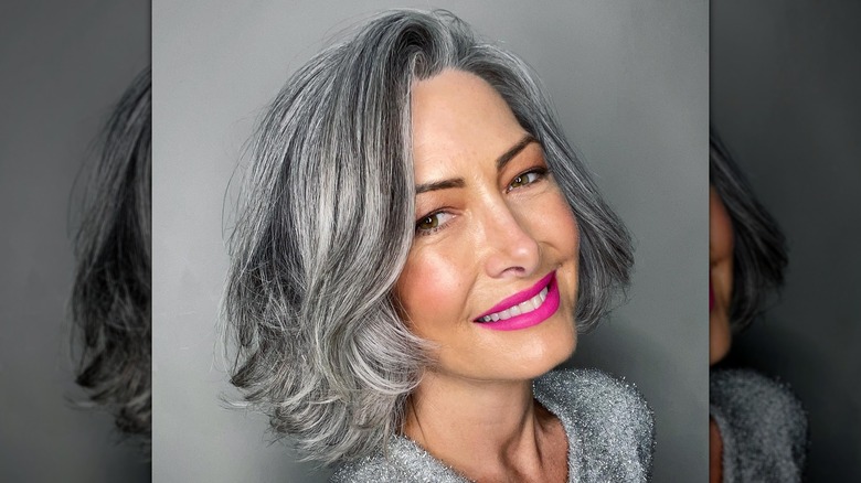 A woman smiling in front of a gray background with bright pink lipstick and a gray bob