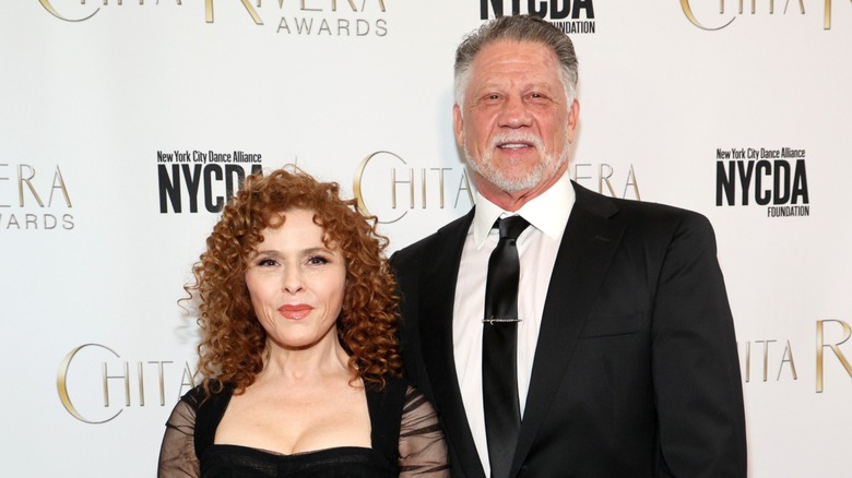 Bernadette Peters in a black dress with sheer sleeves with Tom Sorce on the red carpet in front of a white background in 2024.