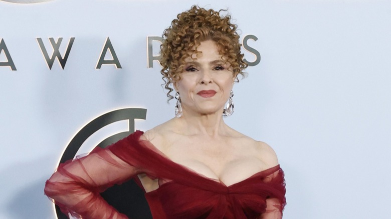 Bernadette Peters posing on the red carpet with her hand on her hip in 2026 wearing a red dress with her curly hair up.