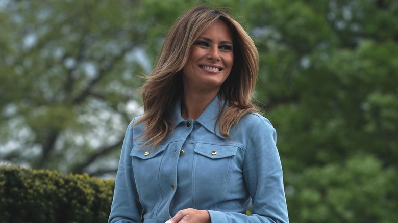 Melania Trump smiling in front of greenery, wearing a blue button-up top