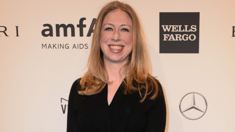 Chelsea Clinton smiling and wearing a black dress on the red carpet while standing in front of a white background with logos