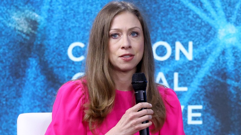 Chelsea Clinton speaking into a microphone on stage in a pink dress