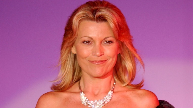 Vanna White smiling and walking in front of a purple background wearing an obvious necklace