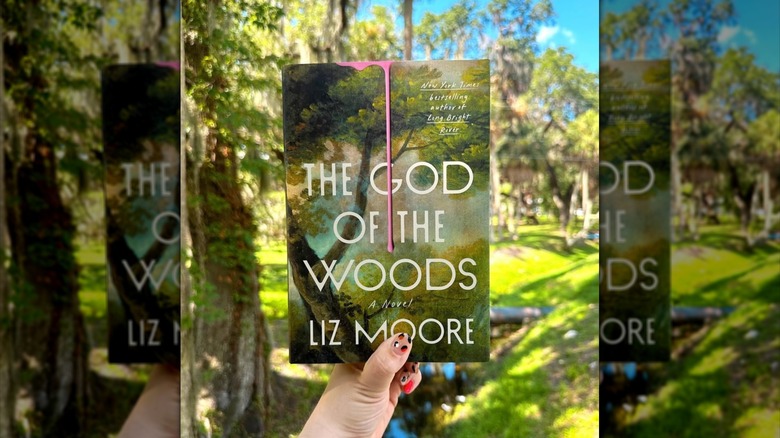 A hand holding a copy of "The God of the Woods" in front of trees