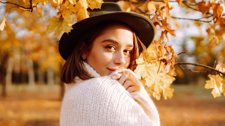 A woman standing under orange leaves with a bob haircut while wearing a white sweater and hat
