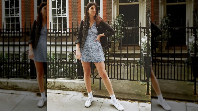 A woman in a short dress and a brown jacket outside wearing Keds