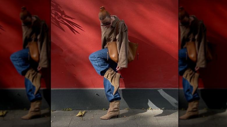 A woman bending down on the street to adjust her suede boot in front of a red wall