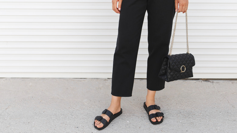 Close-up of person's legs in black pants holding black bag wearing black Birkenstock sandals
