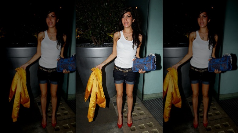 Amy Winehouse poses wearing a white vest and denim mini-shorts, holding a yellow bomber jacket