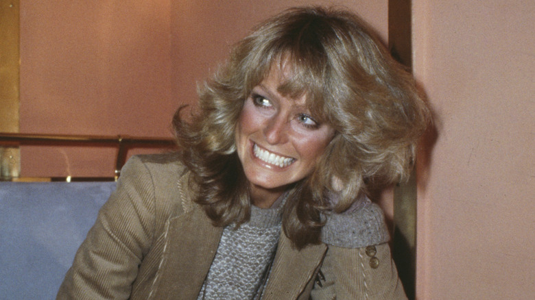 Farrah Fawcett smiling and leaning against a table as her hair is styled into feathered layers