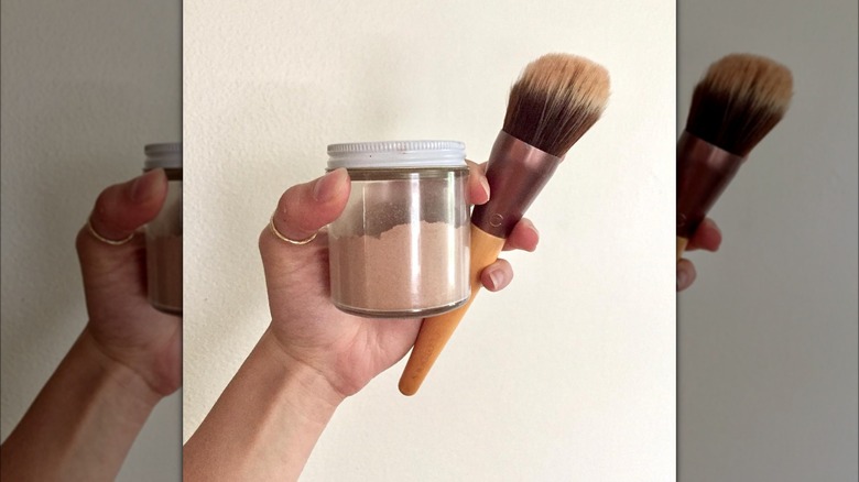 A hand holding up a jar f cocoa powder and a fluffy makeup brush