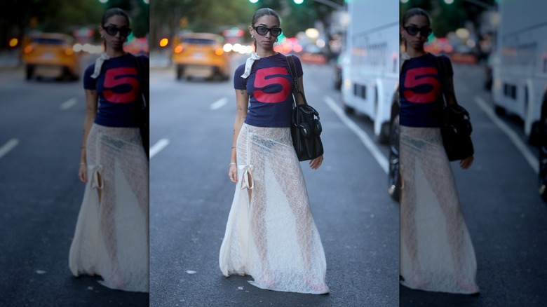 Kayla Curtis-Evans is seen wearing navy blue t-shirt with a large red number five, sheer white lace wrap skirt, pink flats and carrying a black handbag outside of the Elena Velez show, during New York Fashion Week