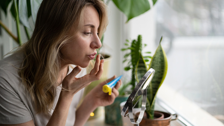 A woman applying lip ointment to her lips with house plants behind her