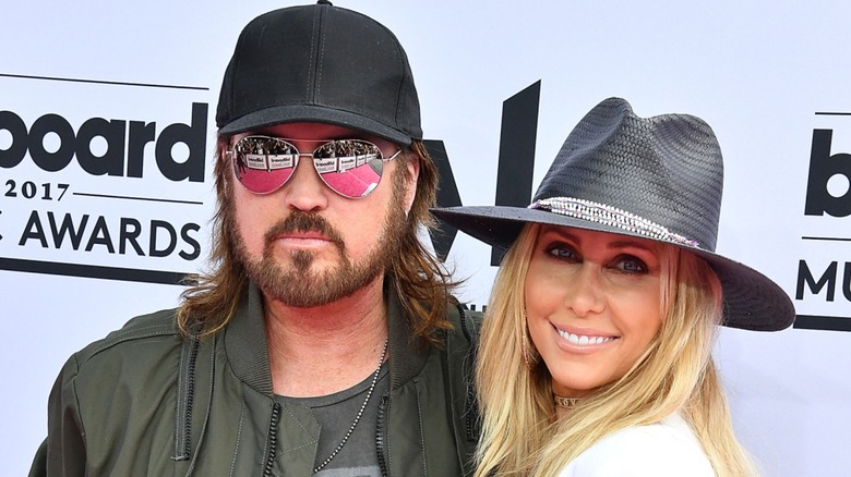 Billy Ray Cyrus in a hat and sunglasses standing alongside Tish Cyrus in a fedora on the red carpet